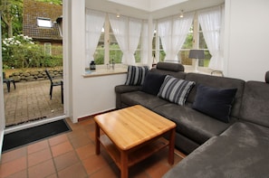 Ein Wohnzimmer mit einer Couch, einem Holztisch und Blick auf einen Garten durch große Fenster.