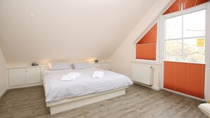 A bedroom with a sloped ceiling, a bed with white linens, a radiator, and orange blinds on a window. - Ferienwohnung mit 3- Zimmern Über ca. 100m² im Obergeschoß und Spitzboden (Wyk auf Föhr)