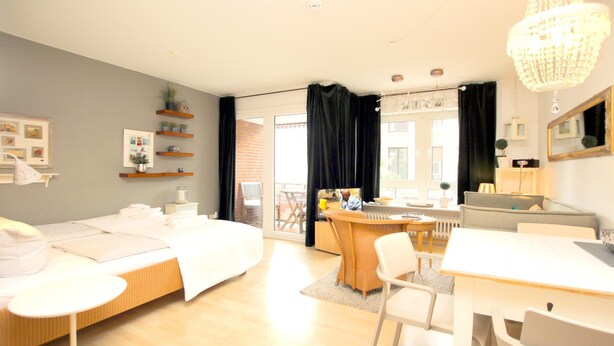 Ett modernt hotellrum med en säng, ett matbord, stolar, en liten köksdel och en balkong med utsikt.