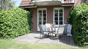 Eine Terrasse mit Tisch und Stühlen, ein Ziegelhaus mit rotem Dach und ein hoher Hecken.