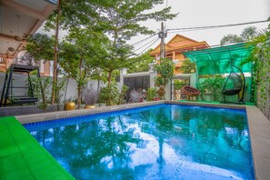 A swimming pool with a tiled edge, surrounded by greenery and a house with a covered patio. - Angkor Rithy Residence (Siem Reap)