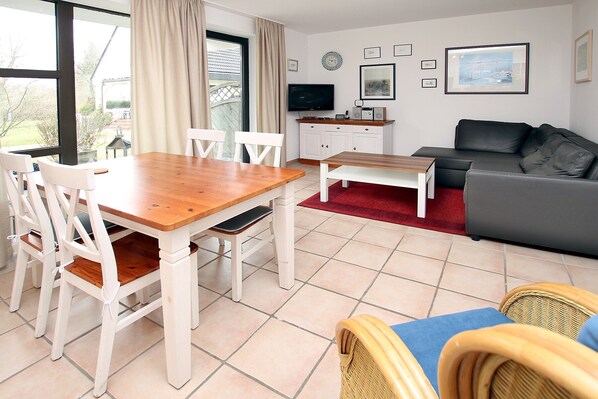Un salon comprenant une table à manger en bois, des chaises blanches, un canapé gris, des fauteuils en osier, un téléviseur et un tapis rouge.