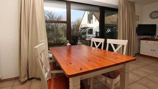 A dining area with a wooden table and chairs, a TV, and a view of a house through large windows.