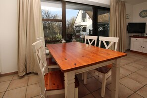 Un coin repas avec une table et des chaises en bois, un téléviseur et une vue sur une maison, visible à travers de grandes fenêtres.