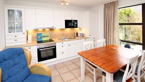 A compact kitchen with white cabinets, a wooden dining table, wicker chairs, and a view through a large window.