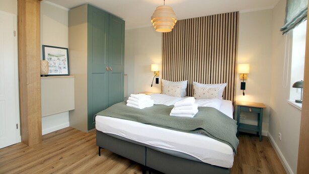 A neatly arranged bedroom with a bed, bedside lamps, a hanging light fixture, a wooden floor, and a wall-mounted cabinet.