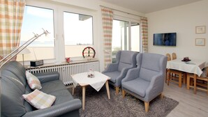 Un salón con un sofá gris, dos sillones, una mesita, un radiador y un televisor en la pared.