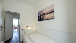 Una habitación pequeña con cama individual, una cama blanca, una mesita de noche, una lámpara de pared y un cuadro de un atardecer sobre un muelle.