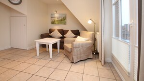 Ein Wohnzimmer mit einer Couch, einem Sessel und einem Tisch. An der Wand hängt eine Uhr, ein Gemälde und ein Fenster mit Jalousien.