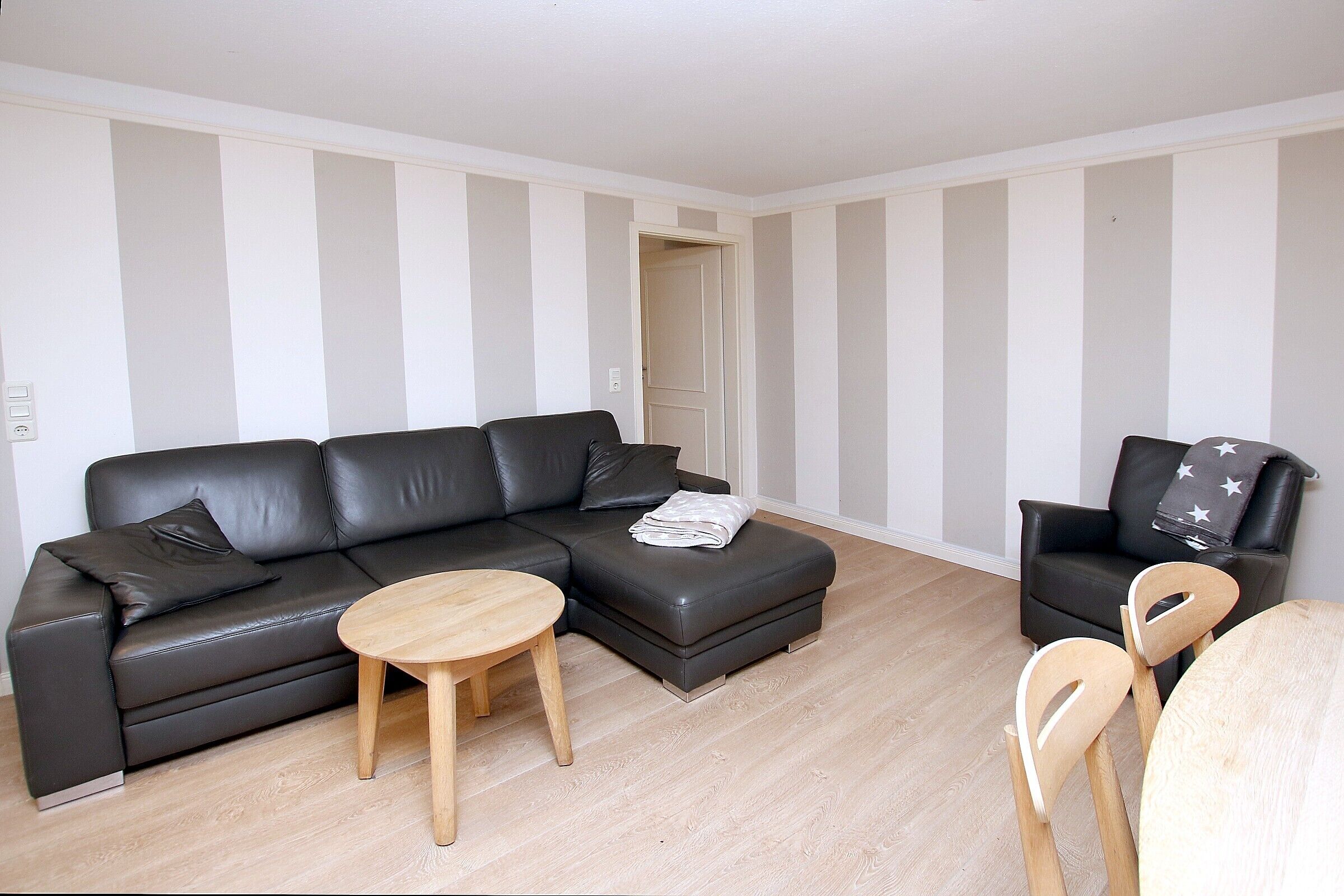 Uma sala de estar moderna com um sofá de couro preto, uma mesa de centro redonda de madeira e uma poltrona preta com um cobertor estampado.