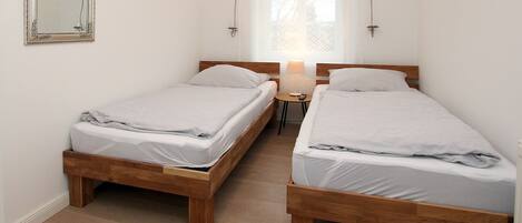 A bedroom with two wooden beds, a bedside table with a lamp, a mirror on the wall, and a window with a lace curtain.