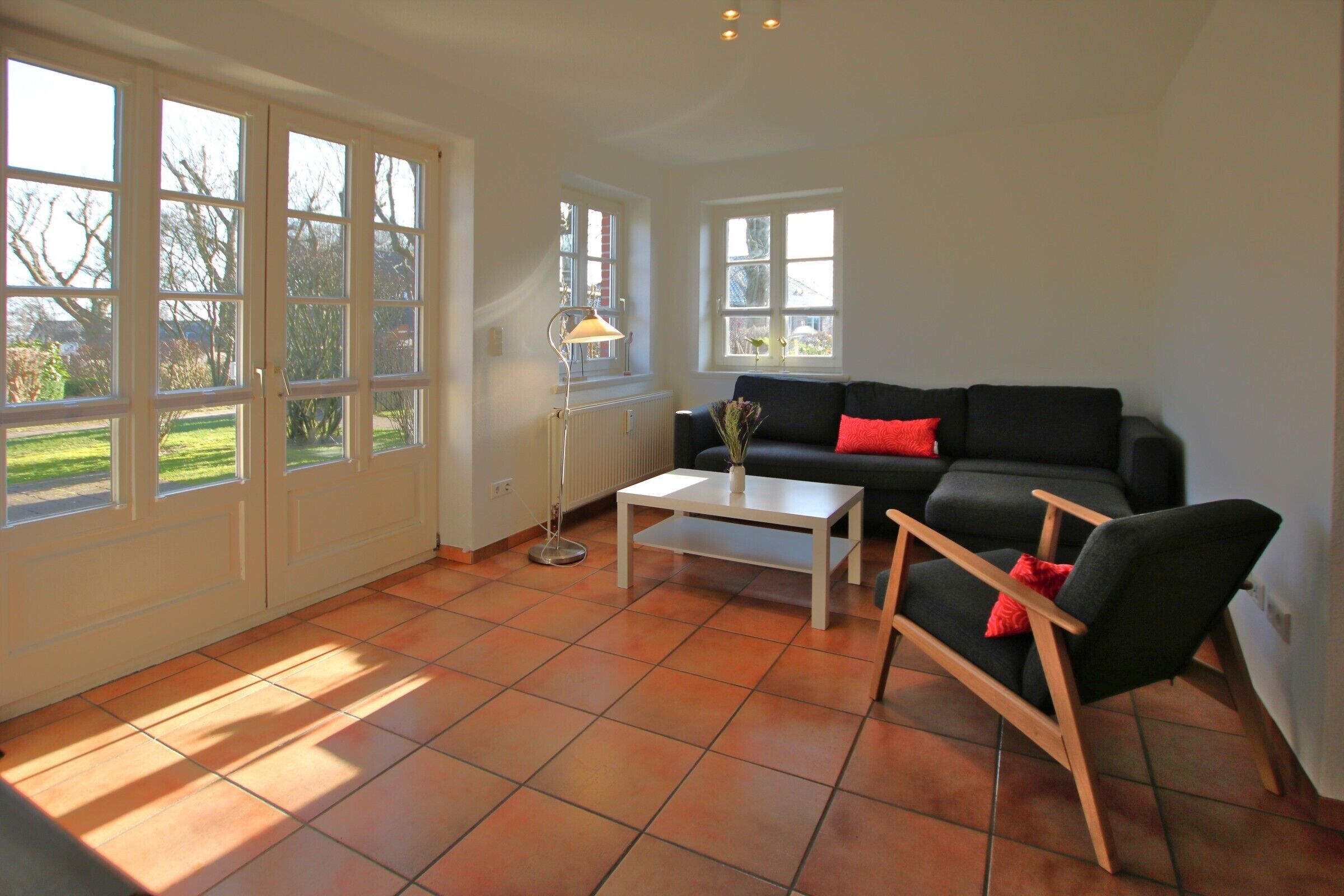 A spacious living room with a black sofa, a white coffee table, and a black armchair. There are large windows allowing natural light and views of the outdoors.