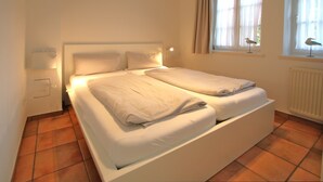 A double bed with white linens in a room with tiled flooring and a window with curtains. - Ferienwohnung mit 3- Zimmern auf ca. 49m² (Wyk auf Föhr)