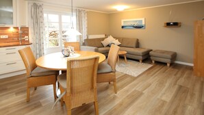 A modern living room with a dining table, wicker chairs, a sofa, and a view of the outdoors through large windows. - Ferienwohnung mit 3- Zimmern auf ca. 50m² (Wyk auf Föhr)