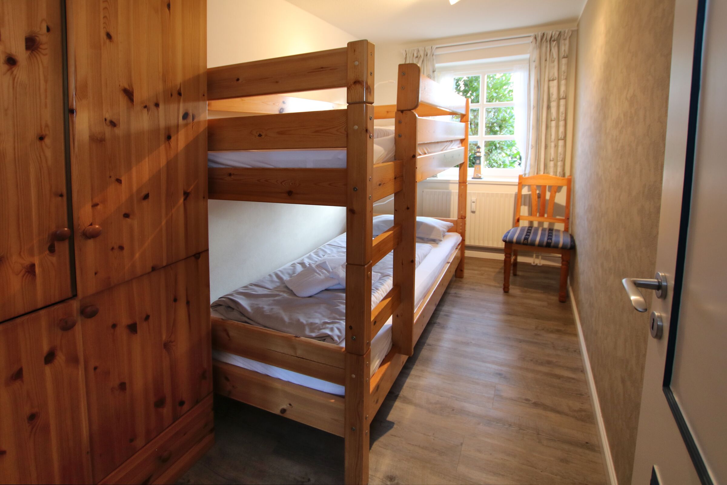 A bunk bed room with a wooden wardrobe and a chair by the window.