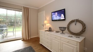 A living room with a television on the wall, a white cabinet, a wicker wreath, and a view of the outdoors through a sliding glass door. - Ferienwohnung mit 3 Zimmern, Sehr Komfortabel und Komplett Renoviert auf 58 m² (Wyk auf Föhr)