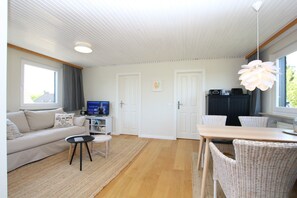 A modern living room with a sofa, a small table, and a dining area with chairs. There is a television on a stand, a bookshelf, and a kitchen area with cabinets and a countertop.