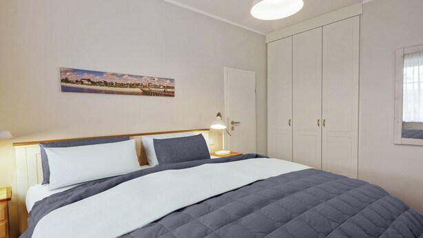 Una cama bien hecha con colcha gris, una lámpara de noche y un cuadro en la pared.