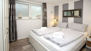 Un lit double avec du linge de lit blanc, une table de chevet avec une lampe, une fenêtre avec des rideaux et une petite étagère avec des objets décoratifs.