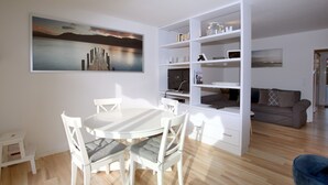 Ein modernes Wohnzimmer mit einem weißen Esstisch und Stühlen, einem Bücherregal, einem Sofa und einem Gemälde an der Wand.