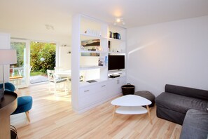 A modern living room with a white entertainment unit, a grey sofa, a white coffee table, and a dining area with a white table and blue chairs. - Ferienwhg. in Wyk mit 3- Zimmern, ca. 58 m² im EG mit Südterrasse und Garten (Wyk auf Föhr)