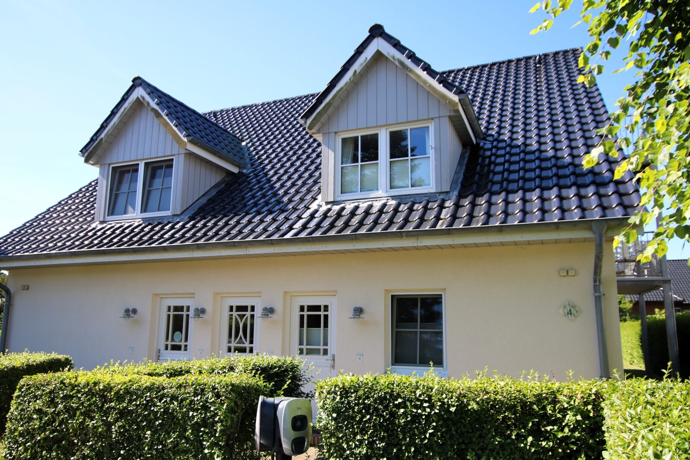 Une maison à deux étages, avec un toit recouvert de tuiles, de nombreuses fenêtres et une haie bien entretenue à l’avant.