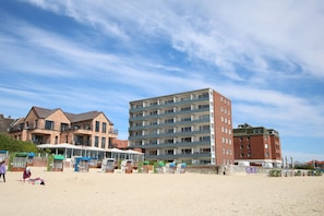 Ein Strand mit einer Reihe von Liegestühlen und Sonnenschirmen, ein großes Backsteinhaus mit mehreren Balkonen und einem klaren Himmel.