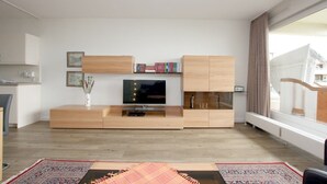 A modern living room with a wooden entertainment unit, a flat-screen TV, and a large window with curtains. - Ferienwhg. Wyk, mit 2 Zimmern, ca. 52 m² im 1.og mit Meerblick auf die Halligen (Wyk auf Föhr)