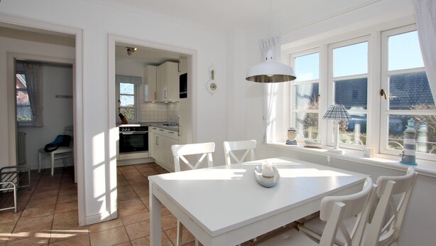 A bright kitchen with white cabinets, a dining table with chairs, and a window with curtains. - Reihenhausscheibe mit 3- Zimmern (Wrixum)