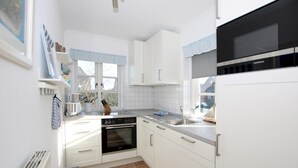 A modern kitchen with white cabinets, a built-in oven, a microwave, and a window with blue curtains. - Reihenhausscheibe mit 3- Zimmern (Wrixum)