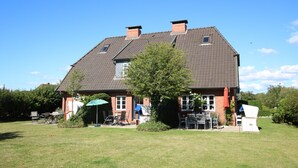 A two-story house with a sloped roof, a chimney, and a small window. There is a lawn with outdoor furniture and a tree in front. - Reihenhausscheibe mit 3- Zimmern (Wrixum)