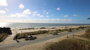 Una carretera costera con una playa al fondo, cielo despejado y algunas personas caminando.