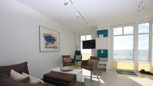 Una sala moderna con un sofá marrón, una mesa de centro y un televisor. Hay un balcón con vista al océano.