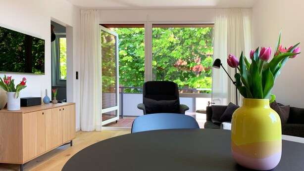 Ein modernes Wohnzimmer mit großem Fenster, einem Holzkabinett, einem schwarzen Sessel und einer gelben Vase mit Tulpen.