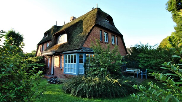 Ein traditionelles Haus mit einem Reetdach, einem Wintergarten und einem Garten mit einer Bank und Stühlen.
