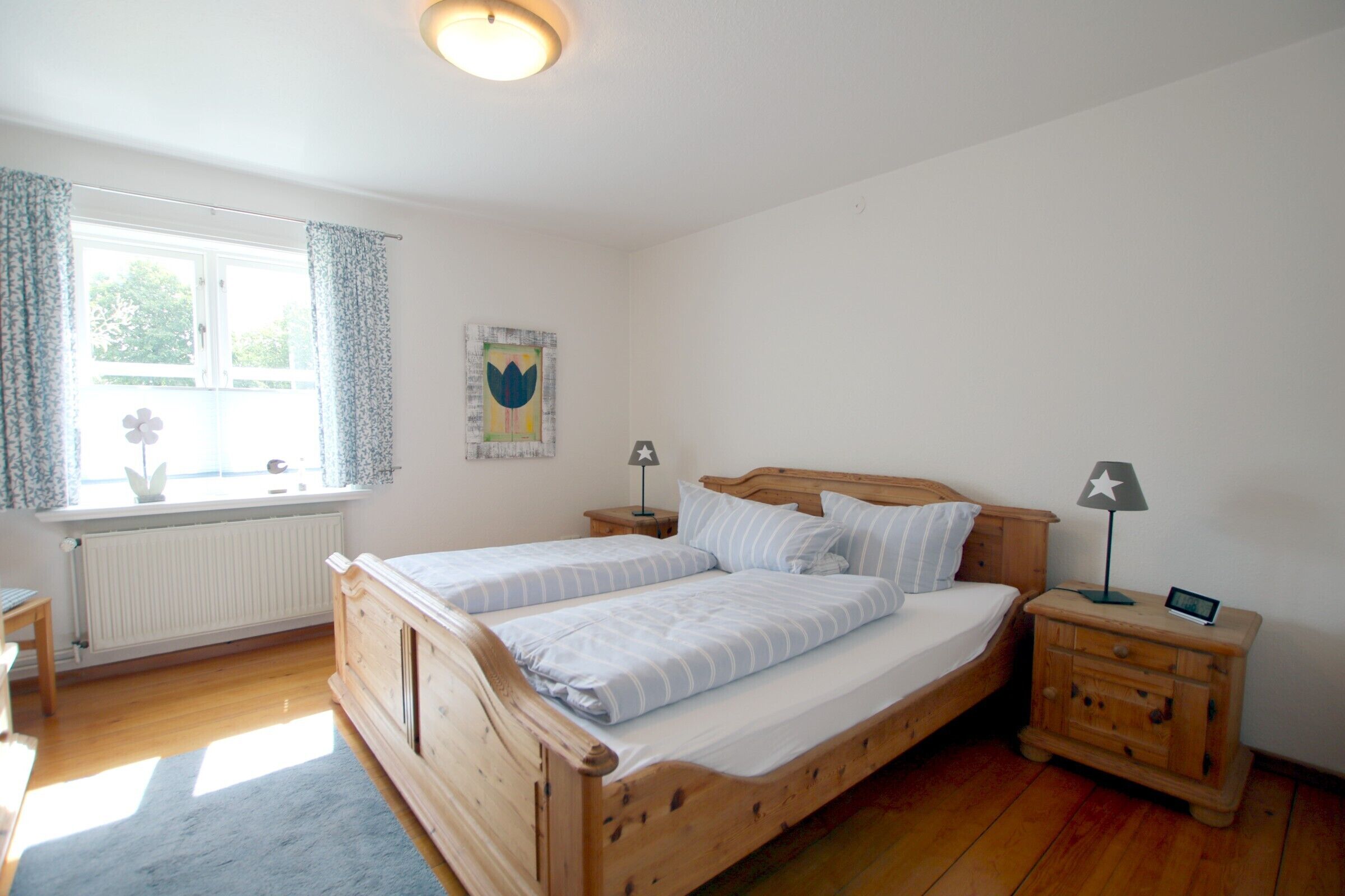 A bedroom with a wooden bed, two bedside tables, a nightstand, a window with curtains, a radiator, and a wall-mounted picture.