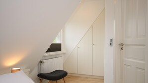 Ein Schlafzimmer mit schrägen Wänden, einem Heizkörper, einem schwarzen Stuhl, einer weißen Tür und einem Fenster mit Blick.