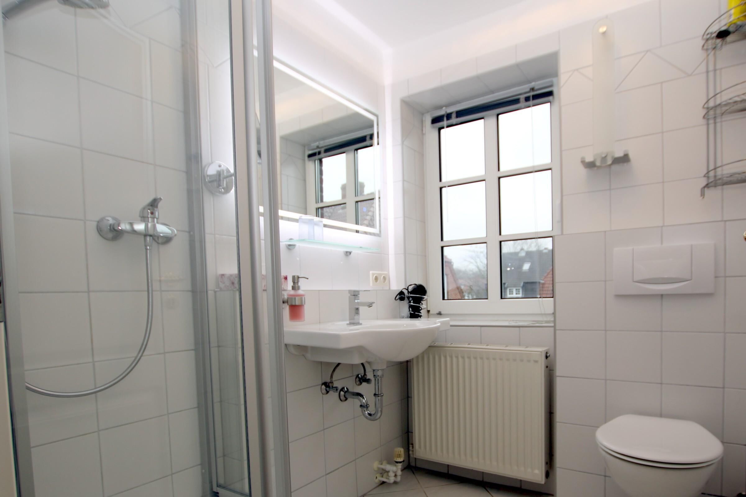 Ein Badezimmer mit Duschkabine aus Glas, Waschbecken in Weiß, einem Fenster und einer Toilette.