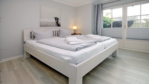 A modern bedroom with a large bed, a bedside table with a lamp, and a view through a window with curtains.