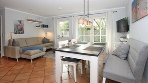 A modern living room with a sofa, dining table, and chairs. There is a TV mounted on the wall and a window with curtains.