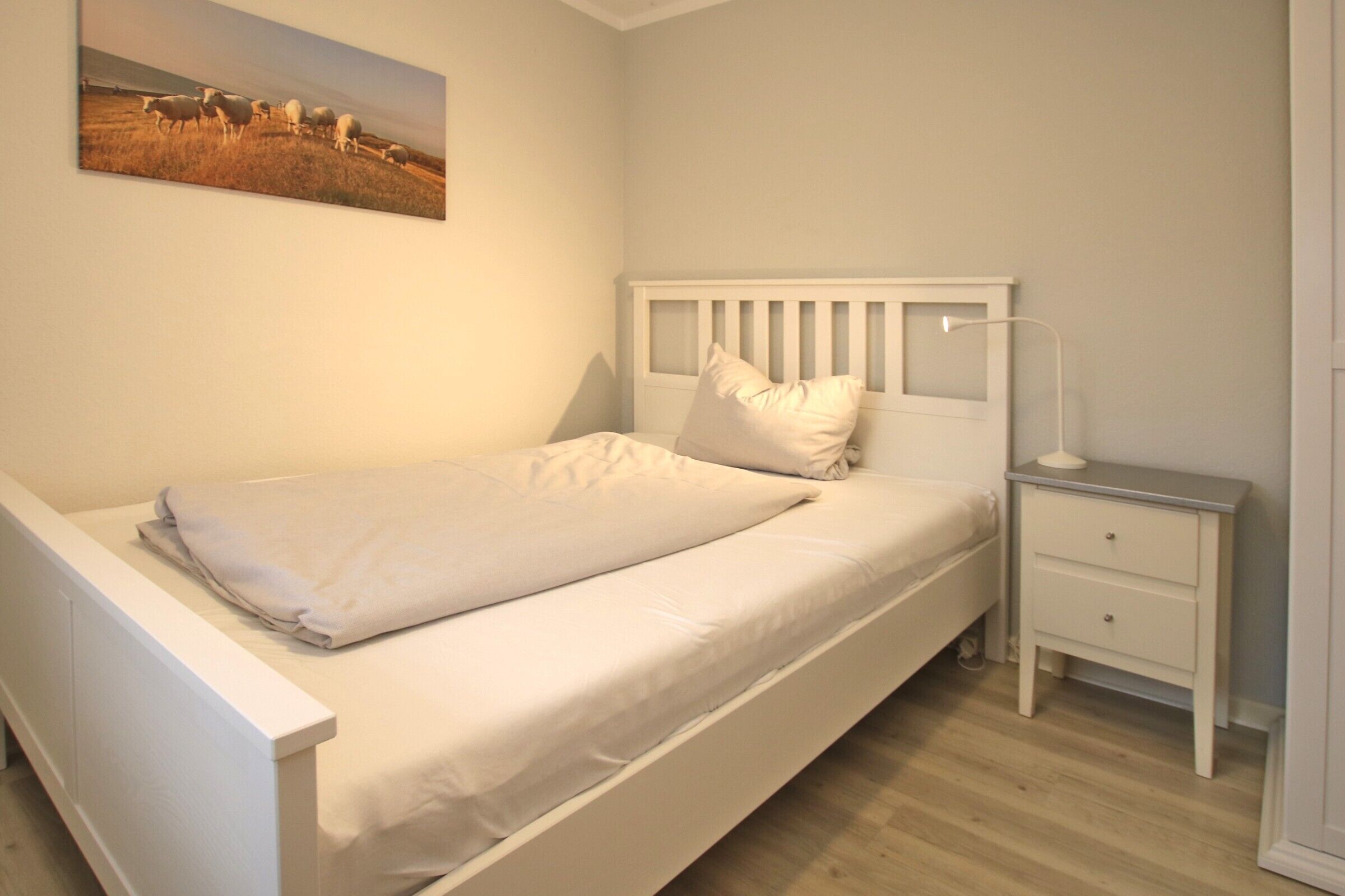 Une chambre à coucher avec un lit blanc, une table de chevet et une commode, ainsi qu’un tableau accroché au mur.