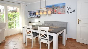 A dining area with a white table and chairs, a television mounted on the wall, and a beach-themed poster.