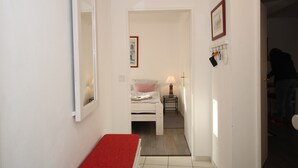 A narrow hallway leading to a bedroom with a bed, a bedside table with a lamp, and a small rug. - Ferienwohnung für 2 Personen auf ca. 44 m² (Wyk auf Föhr)
