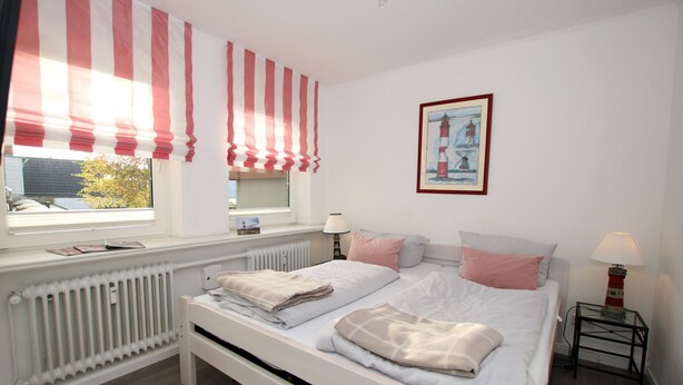 A bedroom with a bed, a window with striped curtains, a radiator, a bedside table with a lamp, and a framed picture on the wall. - Ferienwohnung für 2 Personen auf ca. 44 m² (Wyk auf Föhr)