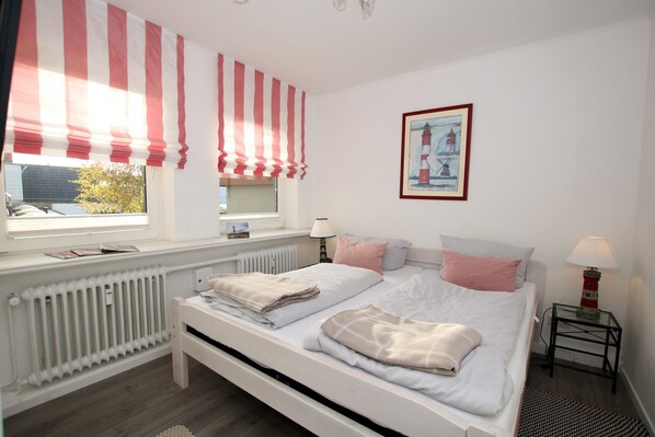 Ein Schlafzimmer mit einem Bett, einem Fenster mit gestreifter Vorhang, einem Heizkörper, einem Nachttisch mit Lampe und einem gerahmten Bild an der Wand.