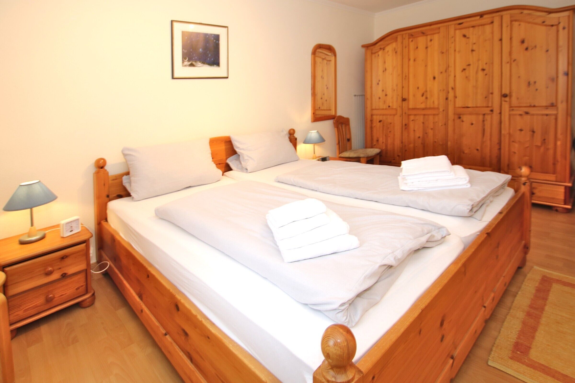 A double bed with white linens and towels, a wooden nightstand with a lamp, a wooden wardrobe, and a framed picture on the wall.