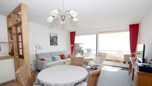 Ein stilvolles Wohnzimmer mit einem runden Esstisch, Korbstühlen, einer Couch und einem großen Fenster mit roten Vorhängen.