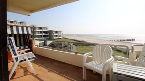 Ein Balkon mit weißen Stühlen und einem Tisch, der Blick auf einen Strand und ein Gebäude mit Balkonen gewährt.