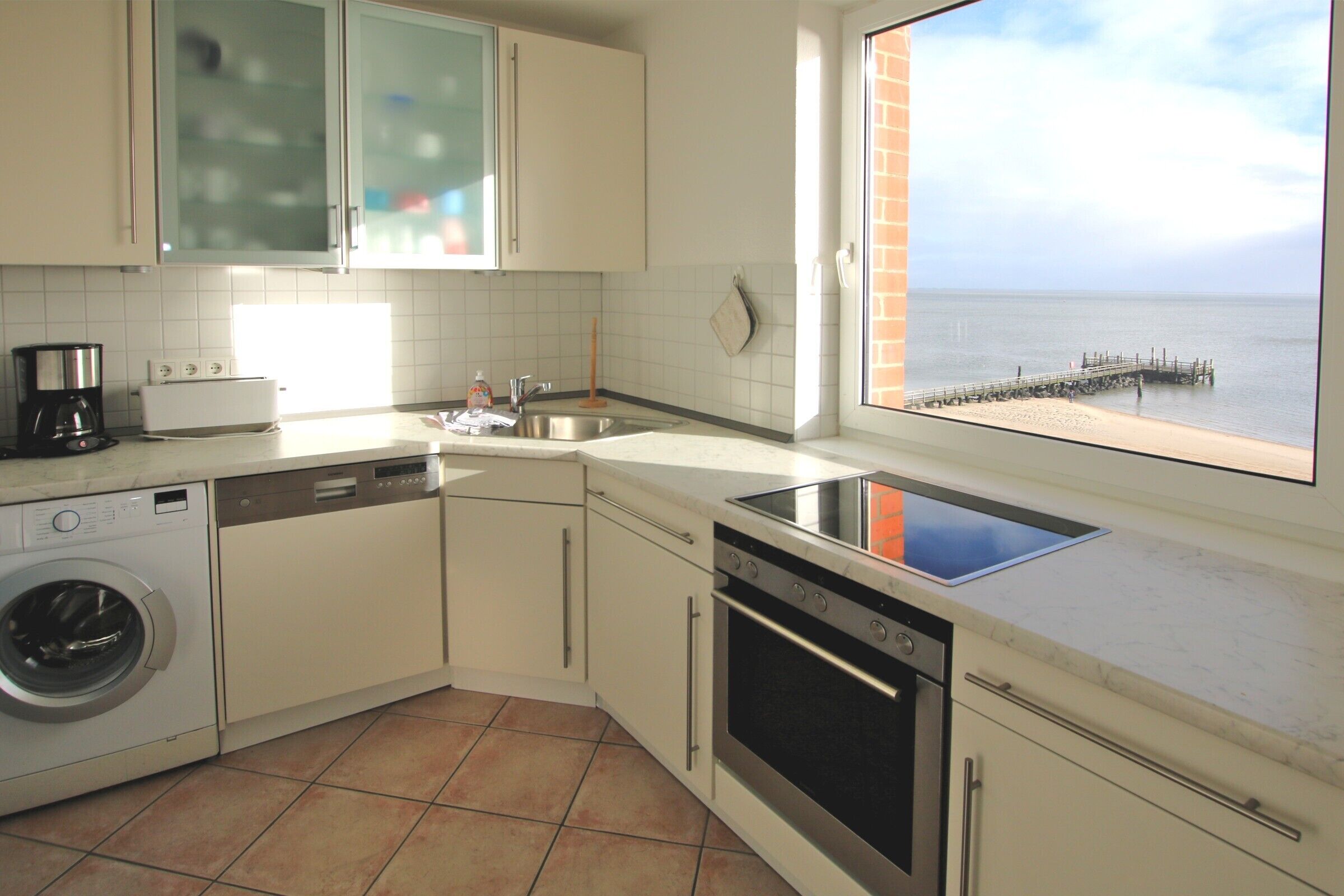 Een keuken met een wasmachine, vaatwasser, fornuis en uitzicht op zee door een raam.