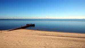 Ein Strand mit einem hölzernen Steg, der sich über ruhiges Wasser erstreckt, einem sandigen Ufer und einem wolkenlosen Himmel.